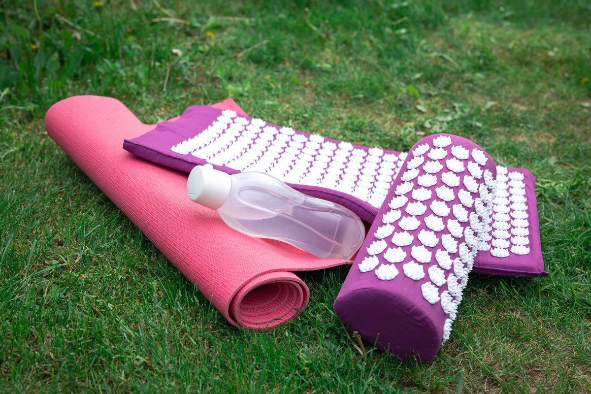 Akkupressur-Set mit lila Matratze, Nackenrolle und Yoga-Matte auf grünem Rasen.