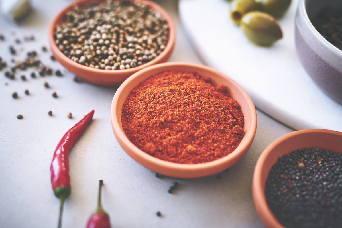 Verschiedene Bio-Gewürze und rote Chilischoten in Tonschalen auf hellem Untergrund.