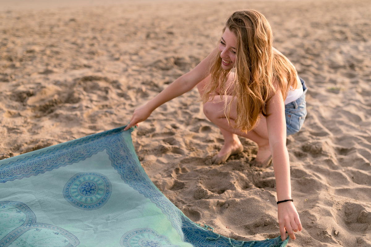 Lächelnde junge Frau breitet ein blaues Yoga-Tuch am Sandstrand aus.