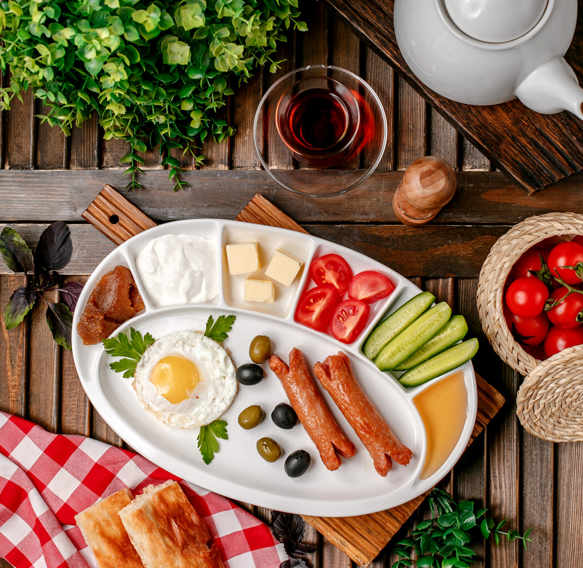Frühstücksteller mit Spiegelei, Würstchen, Tomaten, Gurken, Käse und Oliven auf Holztisch.