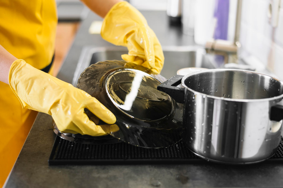 Person mit gelben Handschuhen spült schwarze Teller und einen Edelstahltopf in der Küche.