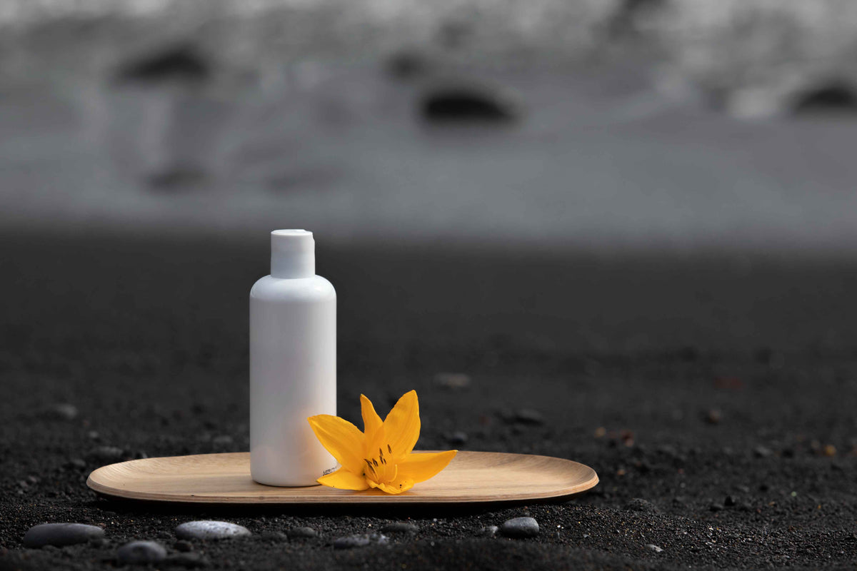 Weiße Bio-Kosmetikflasche mit gelber Blüte auf Holzteller am schwarzen Sandstrand.