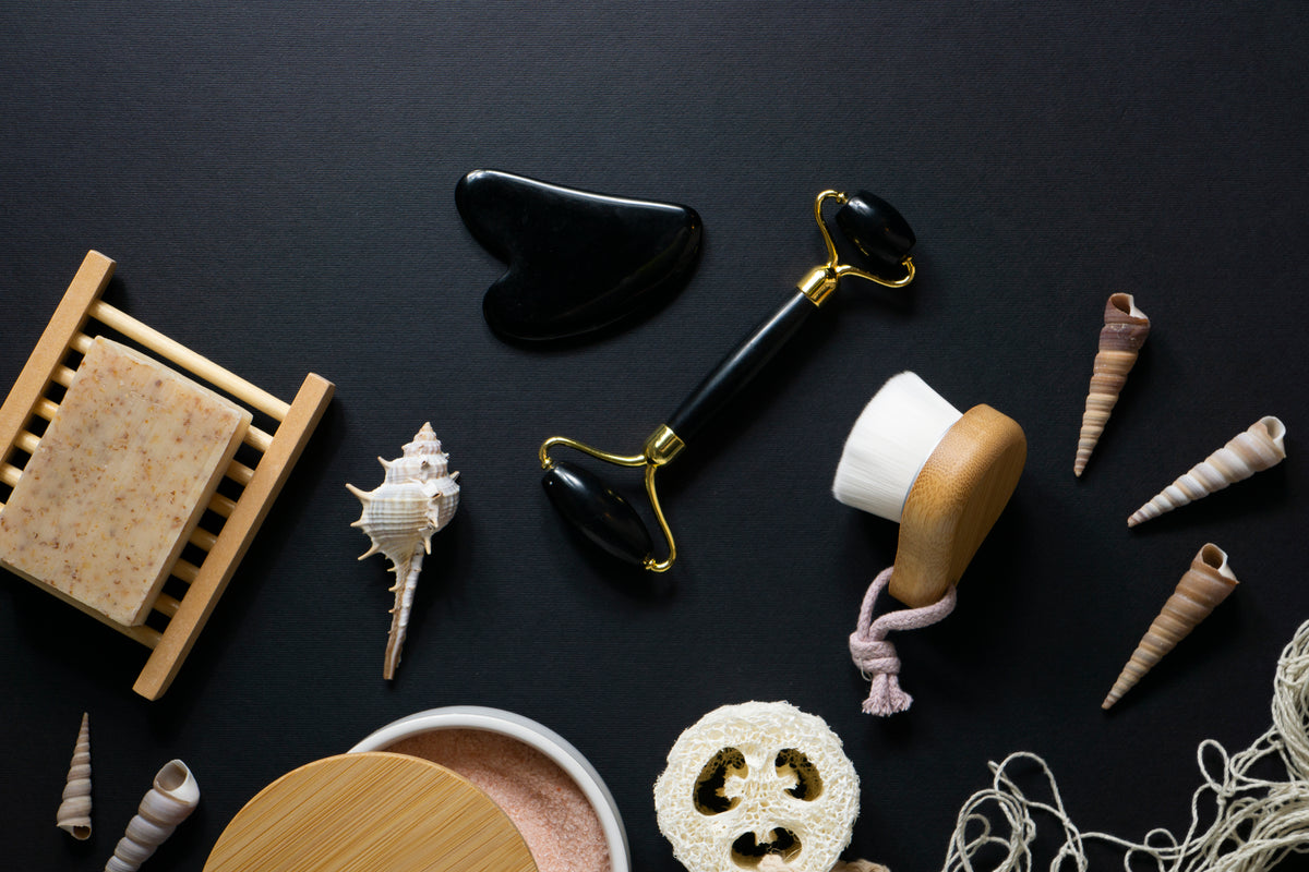 Bio-Kosmetik Zubehör: Schwarzer Gua Sha Stein, Jade-Roller, Naturseife und Muscheln auf schwarzem Grund.