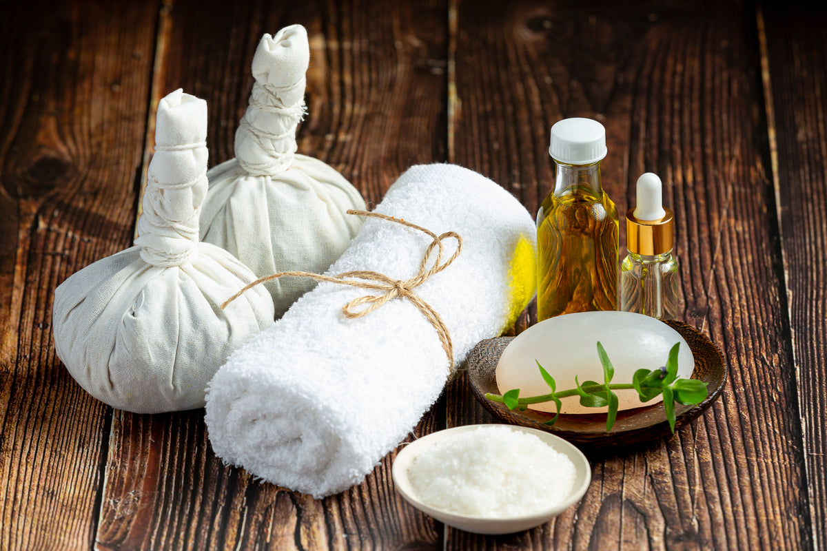 Wellness-Set mit Kräutersäckchen, weißem Handtuch, Massageöl und Naturseife auf Holzuntergrund.