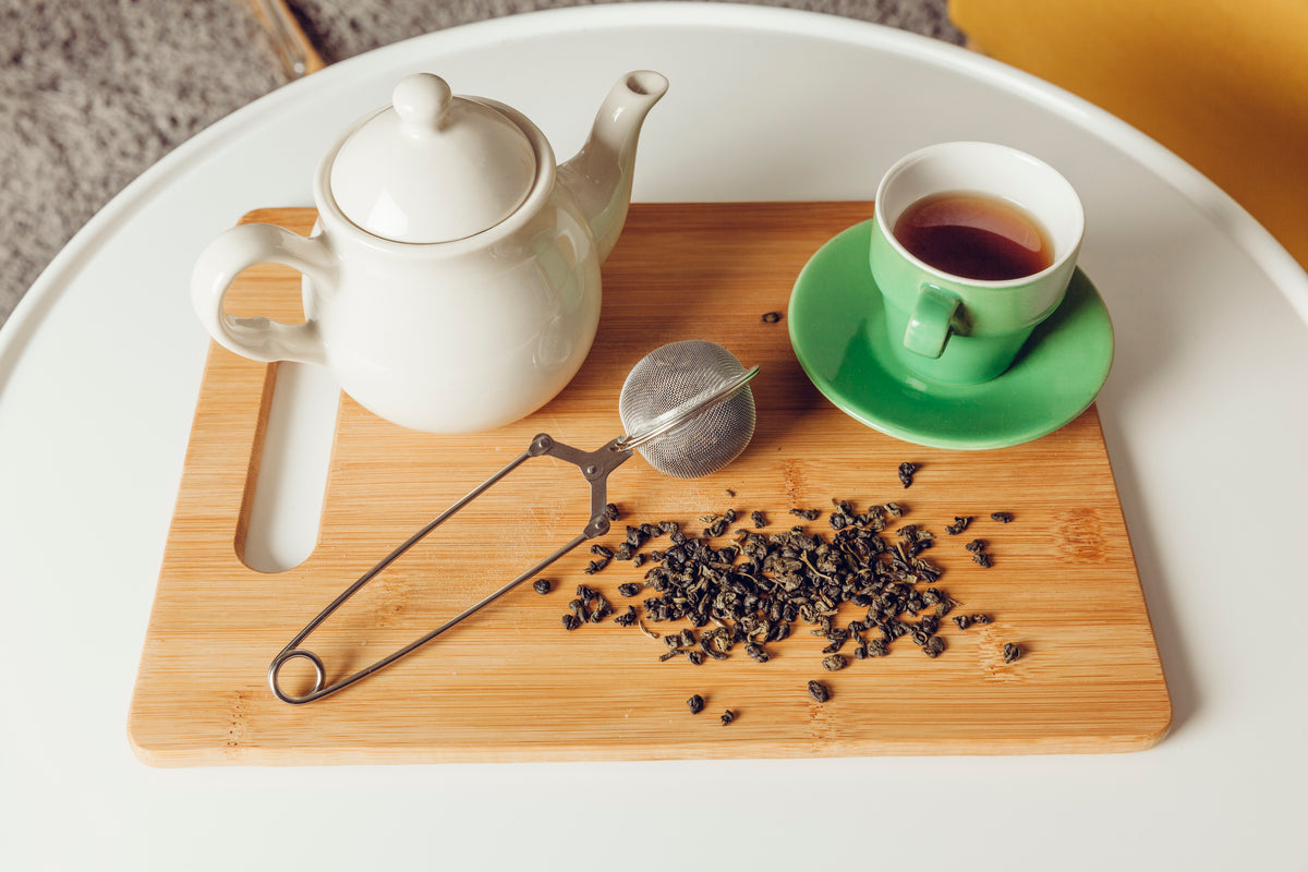 Weiße Teekanne, grüne Tasse mit Tee und lose Teeblätter auf einem Holzbrett.
