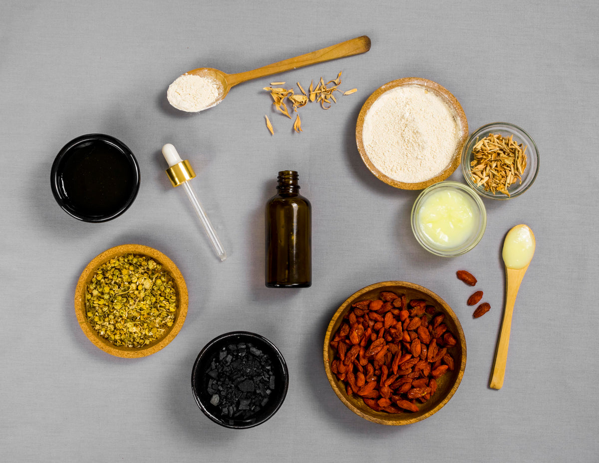 Verschiedene Bio-Kosmetik-Zutaten, Kräuter, Pulver und ätherische Öle in Schalen auf grauem Grund.