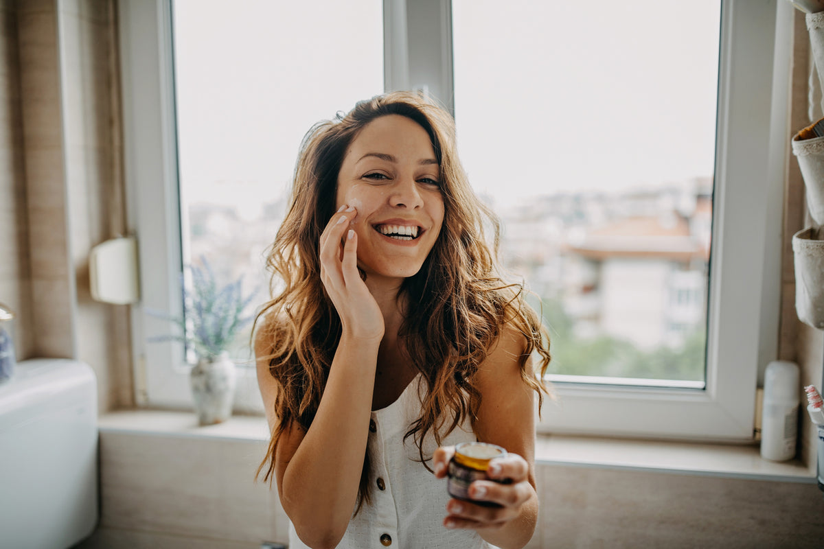 Eine frau mit langen braunen welligen haaren hält ein kleines glas creme in der hand natürliche gesichtspflege rose kumkumadi mana kendra