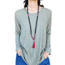 Eine Person trägt eine lange schwarz-rosa Mala-Kette mit silbernem Buddha-Anhänger und roter Quaste zu einem grünen, strukturierten Oberteil und blauen Jeans.