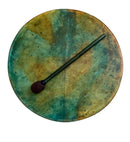 Eine Draufsicht auf eine runde Handtrommel (Rahmen- oder Schamanentrommel) mit einem bemalten Fell in gesprenkelten Gelb-, Orange- und Tiefgrün-/Petroltönen, wobei ein dunkler Holz-Schlägel diagonal auf dem Trommelfell liegt.