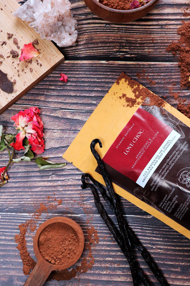 Aufnahme von oben von einer Packung „Love Choc Adaptogenic Cacao Elixir“, die diagonal auf einer dunklen Holzoberfläche platziert ist. Sie ist umgeben von Kakaopulver, einem Holzlöffel mit Pulver, drei dunklen Vanilleschoten, einer roten Rose, Schokolade auf einem Schneidebrett und einem klaren Quarzkristall-Cluster.