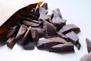 Eine Kraftpapier-Verpackung ist geöffnet und verschüttet dunkle, unregelmäßig geformte Schokoladenstücke auf eine saubere weiße Oberfläche. Etwas Schokoladenstaub ist sichtbar, was auf ein frisches Ausgießen hindeutet.