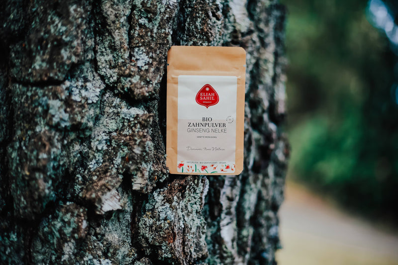 Ein kompostierbarer brauner Papierbeutel mit „Eliah Sahil Bio Zahn pulver Ginseng Nelke“ liegt vertikal an der dunklen, strukturierten Rinde eines Baumes, mit einem unscharfen natürlichen Hintergrund.
