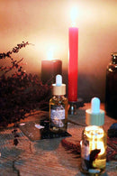 Eine Ritual-Altarszene mit zwei Pipettenflaschen Ritualöl, einer "Lilith"-Orakelkarte und roten Trockenblumen auf einer Holzoberfläche. Im Hintergrund brennen eine hohe rote Kerze und eine schwarze Kerze und tauchen das Arrangement in ein magisches Licht.