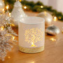 Eine beleuchtete, weiße Keramik-Duftlampe mit leuchtendem Lebensbaum-Ausschnitt vor einem unscharfen Hintergrund aus festlichen silbernen und goldenen Weihnachtsdekorationen und Lichtern.