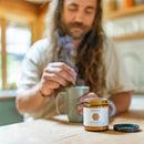 Ein Mann mit langen Haaren und Bart rührt in einer dampfenden Tasse, während im Vordergrund ein offenes Glas Paavani Ashwagandha (3.5 oz / 100g) steht, ein Kräuterergänzungsmittel zur Vitalität und Stresslinderung.