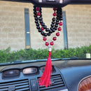 Eine schwarz-rote Mala-Gebetskette mit silbernem Buddha-Anhänger und roter Quaste, die am Rückspiegel eines Autos über dem Armaturenbrett hängt.
