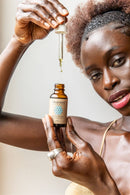 Eine dunkelhäutige Frau hält eine Flasche Paavani Ayurveda Pitta Serum hoch, wobei ein Tropfen des Öls aus der Pipette fällt.