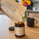 Person, die Paavani Ayurveda Golden Milk (Kurkumapulver) aus einem Beutel in ein Glasgefäß auf einem Holztisch füllt.