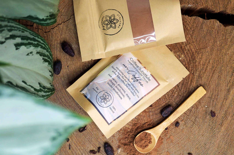 Zwei „Kaffee-Ersatz“-Superfood-Pulvermischungsbeutel liegen auf einem großen, strukturierten Baumstamm. Ein lebhaftes grün-weiß gesprenkeltes Blatt ist links platziert und verleiht der Komposition eine frische, natürliche Note.