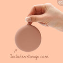 Eine Hand hält eine gefaltete rosa Menstruationsscheibe und weist auf einen "inklusive Aufbewahrungskoffer" auf pfirsichfarbenem Hintergrund hin.