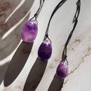 Eine Nahaufnahme von drei polierten Amethyst-Yoni-Eiern in verschiedenen Größen, die an schwarzen Schnüren über einer hellen, marmorierten Oberfläche hängen. Sie liegen teilweise im Schatten und erzeugen starke Schattenlinien über den Kristallen und dem Hintergrund.