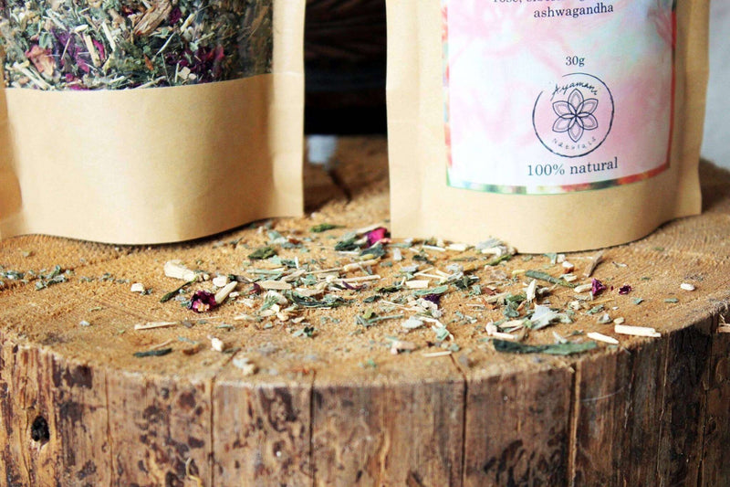 Eine Nahaufnahme einer kleinen Holzschale, gefüllt mit getrockneten grünen Kräutern und leuchtend rosa Rosenblüten, die auf einem strukturierten Baumstumpf ruht. Ein Teil der Kräutermischung ist um die Schale auf der rauen Oberfläche verstreut, was eine organische und natürliche Darstellung schafft.