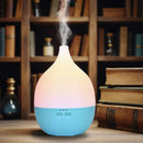 Ein elektrischer, blauer und sanft leuchtender tropfenförmiger Aroma-Diffusor, der Nebel ausstößt und auf einem Holztisch vor einem Bücherregal mit alten Büchern steht.