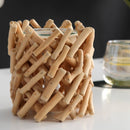 Nahaufnahme eines rustikalen Kerzenhalters aus ineinander verflochtenen, hellen Holzstäbchen, der auf einer weißen Oberfläche neben einem Glas steht.