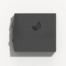 Quadratisches Stück dunkelgrauer Seife mit einem geprägten Blatt-Logo.







