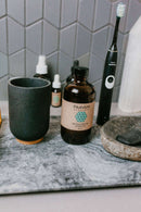 eine Badezimmerablage mit strukturierter grauer Oberfläche und sechseckigen grauen Wandfliesen. Zu den Produkten gehören ein schwarzer Keramikbecher, eine Flasche PAAVANI Oil Pulling Öl, kleinere Flaschen, eine elektrische Zahnbürste und eine Steinschale mit einem schwarzen Stein.