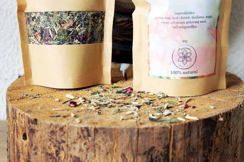 Eine Nahaufnahme einer kleinen Holzschale, gefüllt mit getrockneten grünen Kräutern und leuchtend rosa Rosenblüten, die auf einem strukturierten Baumstumpf ruht. Ein Teil der Kräutermischung ist um die Schale auf der rauen Oberfläche verstreut, was eine organische und natürliche Darstellung schafft.