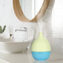 Ein elektrischer, blau-gelb-grüner tropfenförmiger Aroma-Diffusor, der Nebel ausstößt und auf einer Ablage in einem modernen Badezimmer mit Waschbecken und rundem Spiegel steht.