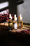 Drei Glasflaschen mit Pipette und Bambusverschluss, gefüllt mit Ritualöl, stehen auf einer Holzoberfläche und werden von Sonnenlicht beleuchtet. Die Flaschen enthalten goldenes Öl und Pflanzenbestandteile. Rote Trockenblumen, ein schwarzer Stein und ein roter Farn sind dekorativ arrangiert, mit einer grauen Wand im Hintergrund.