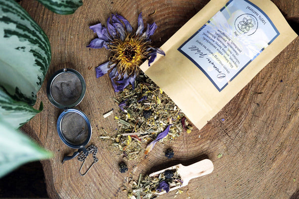 Eine Tüte "Dream Well" Teemischung, eine kleine Holzschaufel, ein Sieb und eine verwelkte Blume liegen auf einem Baumstumpf. Die getrockneten Kräuter, Beeren und Blüten der Teemischung sind auf dem Holz verstreut.