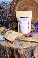 Zwei Teebeutel stehen auf einer Holzoberfläche. Einer ist geöffnet und zeigt lose Teeblätter und getrocknete Blumen, die mit einem Holzgerät aufgesammelt werden. Ein großer geschnitzter Holzteller und getrocknete Blumen sind im Hintergrund zu sehen.