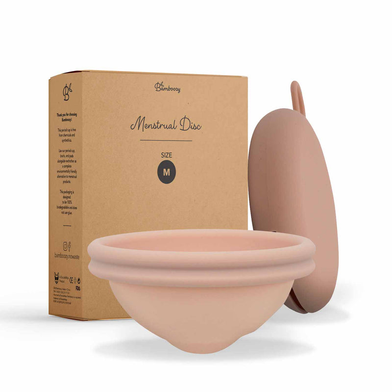 Eine rosa Menstruationsscheibe in Größe M, zusammen mit ihrer braunen Kartonverpackung von Bamboozy, betont die Bezeichnung "Menstruationsscheibe" und umweltfreundliche Merkmale.