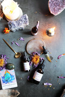 Aufnahme von oben von zwei dunklen Tropfflaschen mit „Blue Lotus Tincture“ auf einer dunklen Oberfläche, umgeben von brennenden Kerzen, Amethystkristallen, getrockneten lila Blüten, einem goldenen Löffel und einer kleinen Orakelkarte.