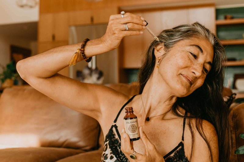 A woman with graying hair applies Paavani Karna Purana Ear Oil to her ear with a dropper, while holding a brown bottle of the oil.
