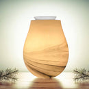 Ein leuchtender, tropfenförmiger elektrischer Wachswärmer mit einem hellen, marmorartigen oder holzmaserungsartigen Muster in Bernstein- und Brauntönen, der auf einer Holzoberfläche mit zwei Kiefernnadelzweigen steht.