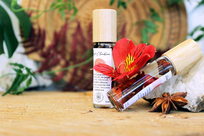 Eine klare Glas-Roll-On-Flasche, beschriftet mit „Root Chakra“, liegt seitlich auf einer rustikalen Holzscheibe und zeigt die botanischen Zutaten und roten Jaspis-Kristallsplitter im Inneren. Dahinter steht ein Bambusdeckel aufrecht, und eine leuchtend rote Lilie setzt rechts einen Farbakzent. Die Szene vermittelt den Eindruck natürlicher, ganzheitlicher Produkte.
