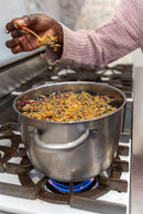 Eine Hand lässt eine Prise getrockneter Kräuter und Rosenblätter in einen großen Topf mit kochendem Wasser auf einem Herd fallen.