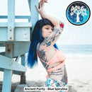 Eine Frau mit leuchtend blauen Haaren und Tattoos, die an einem Strand für Ancient Purity Blaues Spirulina posiert.