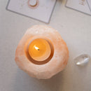 Eine Draufsicht auf einen Himalaya-Salz-Teelichthalter mit einer brennenden, leuchtenden Kerze, der auf einer weißen Oberfläche neben einem klaren Kristall und verschwommenen spirituellen oder Orakelkarten liegt.