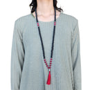 Eine Person trägt eine lange Mala-Gebetskette aus schwarzen und rosa Perlen mit einem silbernen Buddha-Anhänger und einer roten Quaste über einem grauen, strukturierten Oberteil.