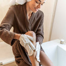 Eine lächelnde Frau in einem braunen Bademantel und einem Handtuch auf dem Kopf sitzt und benutzt weiße Trockenbürsthandschuhe an ihrem Bein.