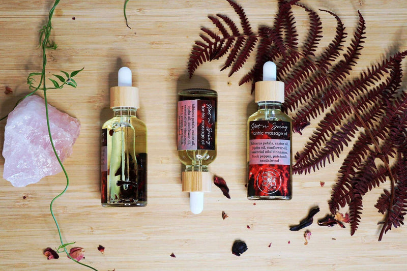 Drei Tropfflaschen "Hot n Spicy Tantric Massage Oil" sind horizontal auf einer Holzoberfläche aufgereiht, verziert mit verstreuten getrockneten roten Blütenblättern und einem leuchtend roten Farn. Ein großer rosa Rosenquarz-Kristall und eine grüne Weinrebe sind links positioniert und tragen zu einer natürlichen und einladenden Darstellung bei.