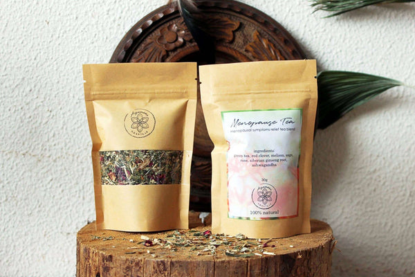  Zwei Pakete Kräutertee liegen auf einer Holzoberfläche. Ein Paket ist mit "Menopause Tea" beschriftet und enthält eine Liste natürlicher Zutaten, während das andere ein dekoratives Design hat.