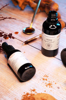 Zwei dunkle Glasflaschen mit Reishi-Flüssigextrakt auf einer Holzoberfläche mit einem goldenen Löffel und Kräuterpulver.