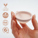 Eine Hand hält eine rosa Menstruationsscheibe und hebt verschiedene Zertifizierungen hervor: tierversuchsfrei und vegan, RoHS-konform, FDA-zugelassen und Qualicert SGS-zertifiziert.