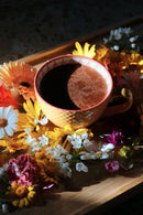 Eine gelbe Tasse mit einem dunklen Getränk und rötlichem Pulver steht mittig auf einem hellen Holztablett, umgeben von einem lebhaften Arrangement bunter Blumen in Orange, Gelb, Weiß und Pink, beleuchtet von einem Sonnenstrahl.